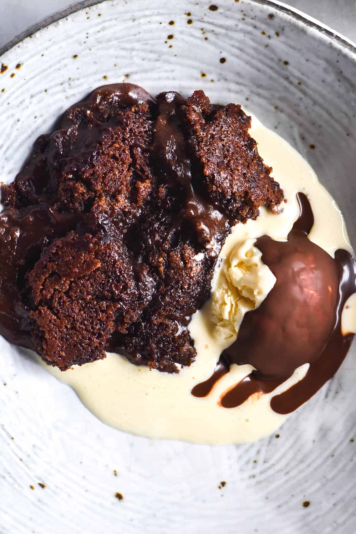 An aerial close up view of a bowl filled with gluten free self saucing chocolate pudding, melting vanilla ice cream and a melted chocolate shell atop the ice cream