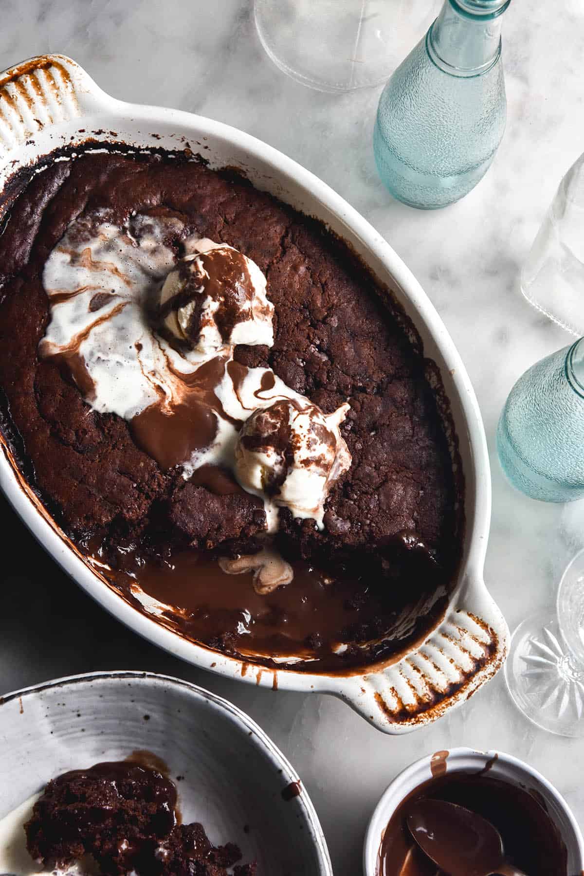 An aerial view of a grain free chocolate self saucing pudding in an oval baking dish. The dish sits on a white marble table surrounded by water glasses and more bowls of pudding. Melted vanilla ice cream and chocolate sauce meld together on the top of the pudding