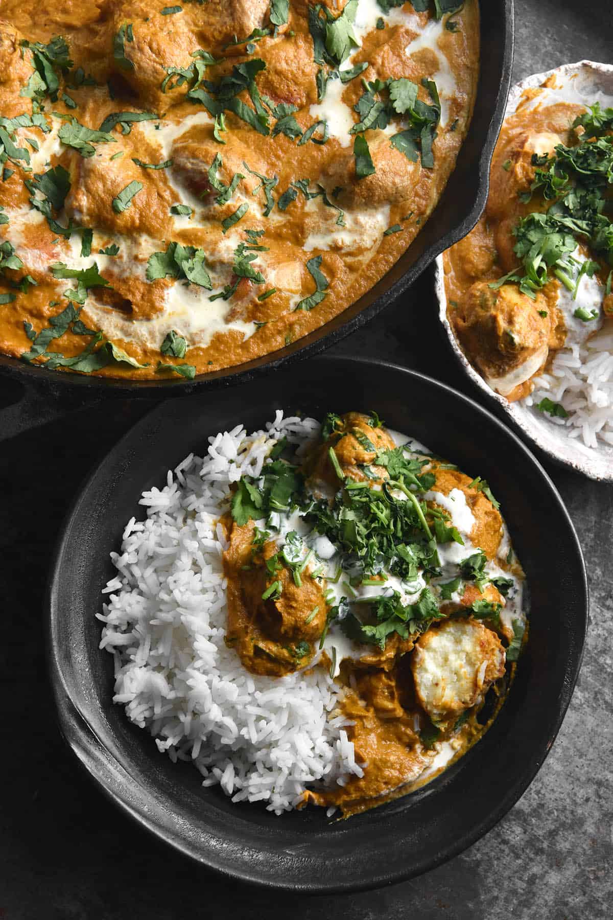 An aerial view of two bowls and a skillet filled with FODMAP friendly Malai Kofta. The skillet kofta is topped with a swirl of cream and coriander. The bowls are filled with white rice accompanying the kofta. The scene is set on a dark grey steel backdrop.