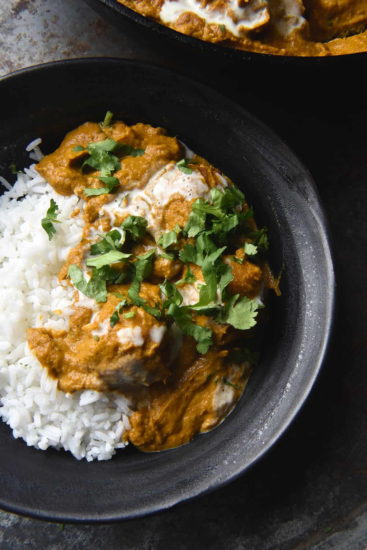 A moody aerial view of a dark grey ceramic bowl filled with rice and FODMAP friendly Malai kofta topped with cream and coriander. A skillet of Malai kofta sits in the top right corner of the image, which is set on a dark grey backdrop.