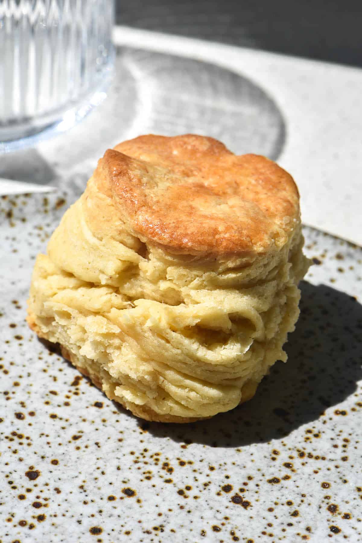 A side on view of a flaky and tall gluten free biscuit on a white speckled ceramic plate. A sunlit glass of water sits in the background.