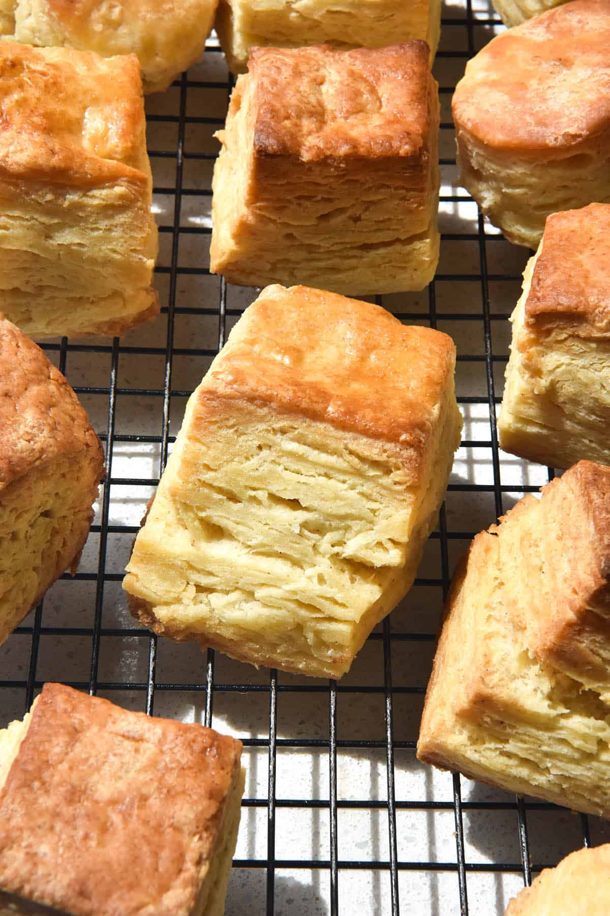 A brightly lit aerial view of gluten free biscuits on a wire baking rack. The biscuits are square, tall and flaky with golden brown tops.