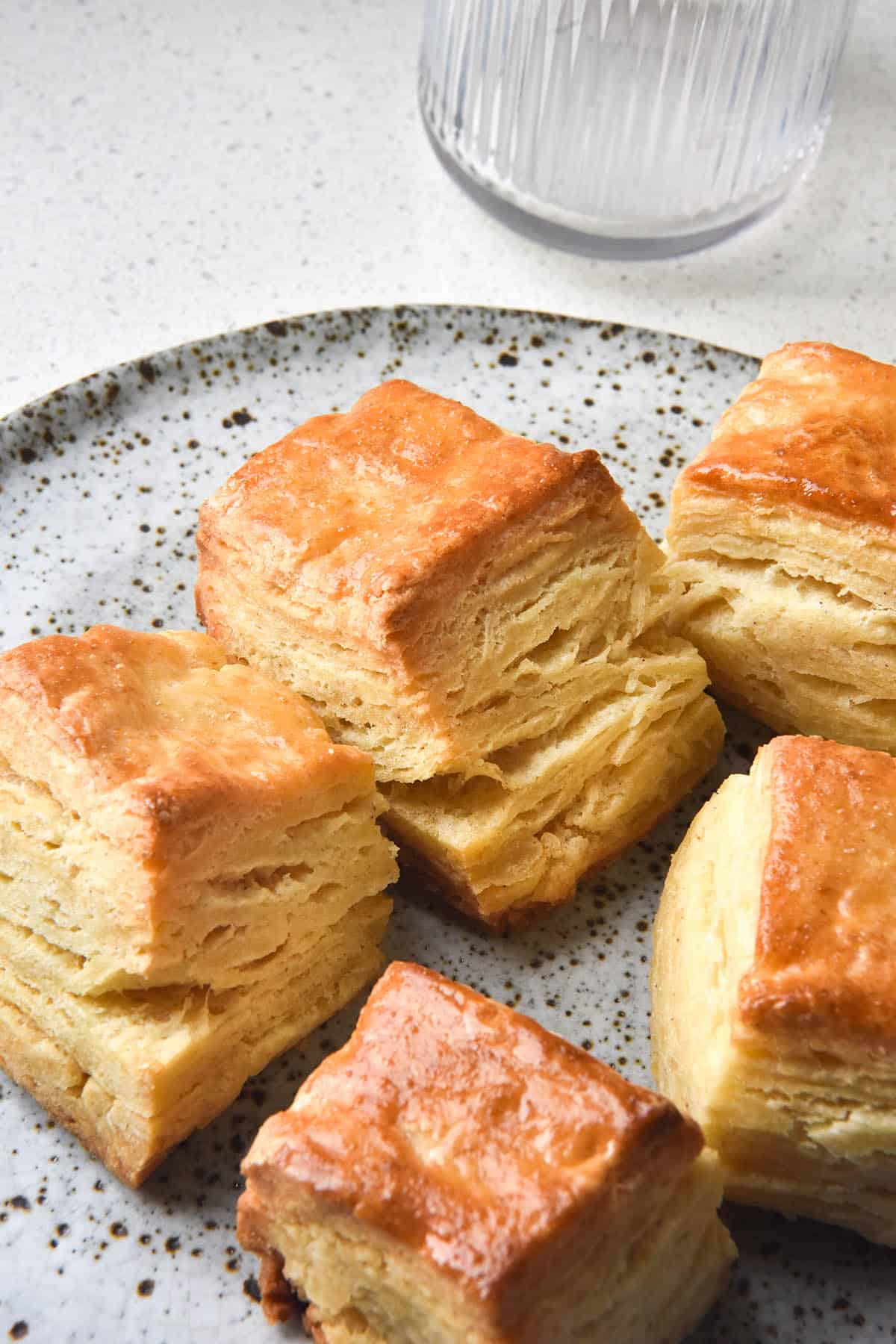 A side on view of five gluten free biscuits on a white speckled ceramic plate. The biscuits are square and have lots of flaky layers. The plate sits on a white benchtop with a textured water glass in the background