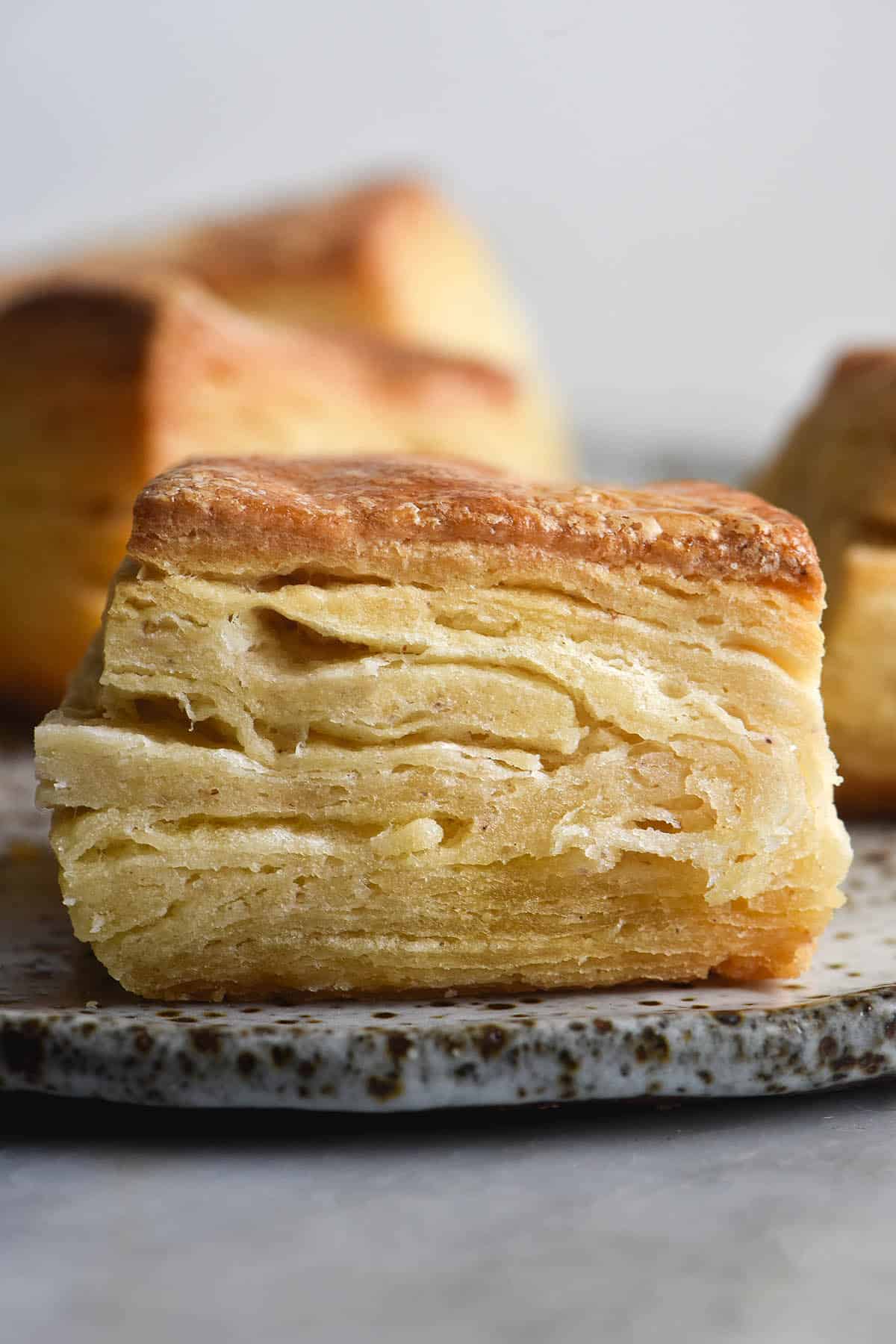 A side on view of a flaky gluten free biscuit on a white speckled ceramic plate. More biscuits sit in the background