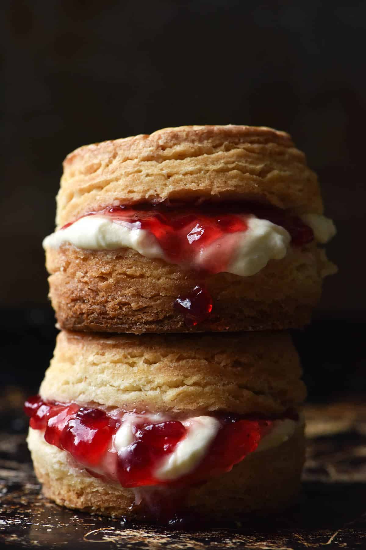 A side on moody image of two gluten free biscuits filled with jam and cream and stacked on top of each other. The biscuits sit on a black tray against a black backdrop. 