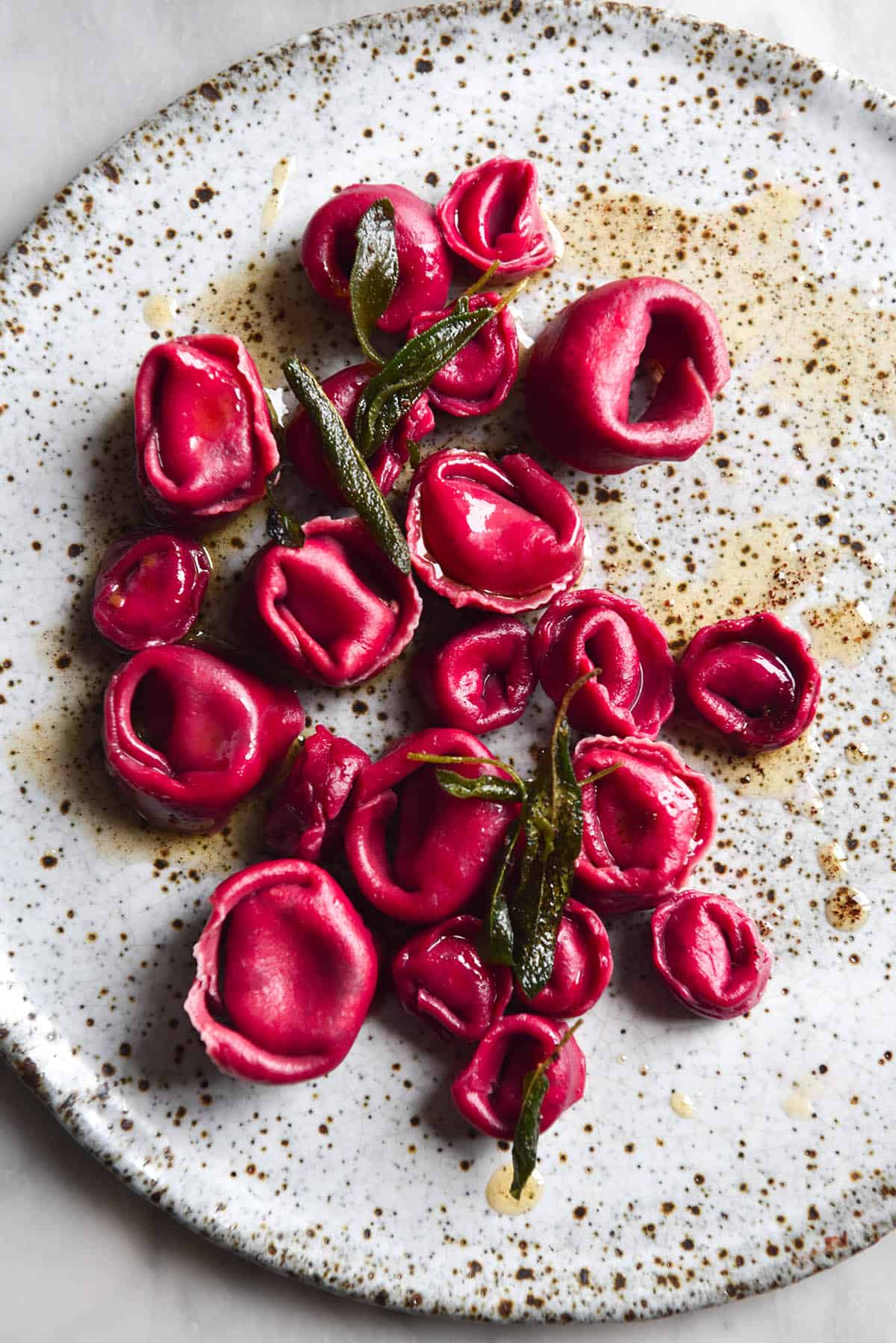 An aerial image of a white ceramic plate topped with gluten free ravioli made from beetroot dough and topped with a brown butter and crispy sage sauce. 