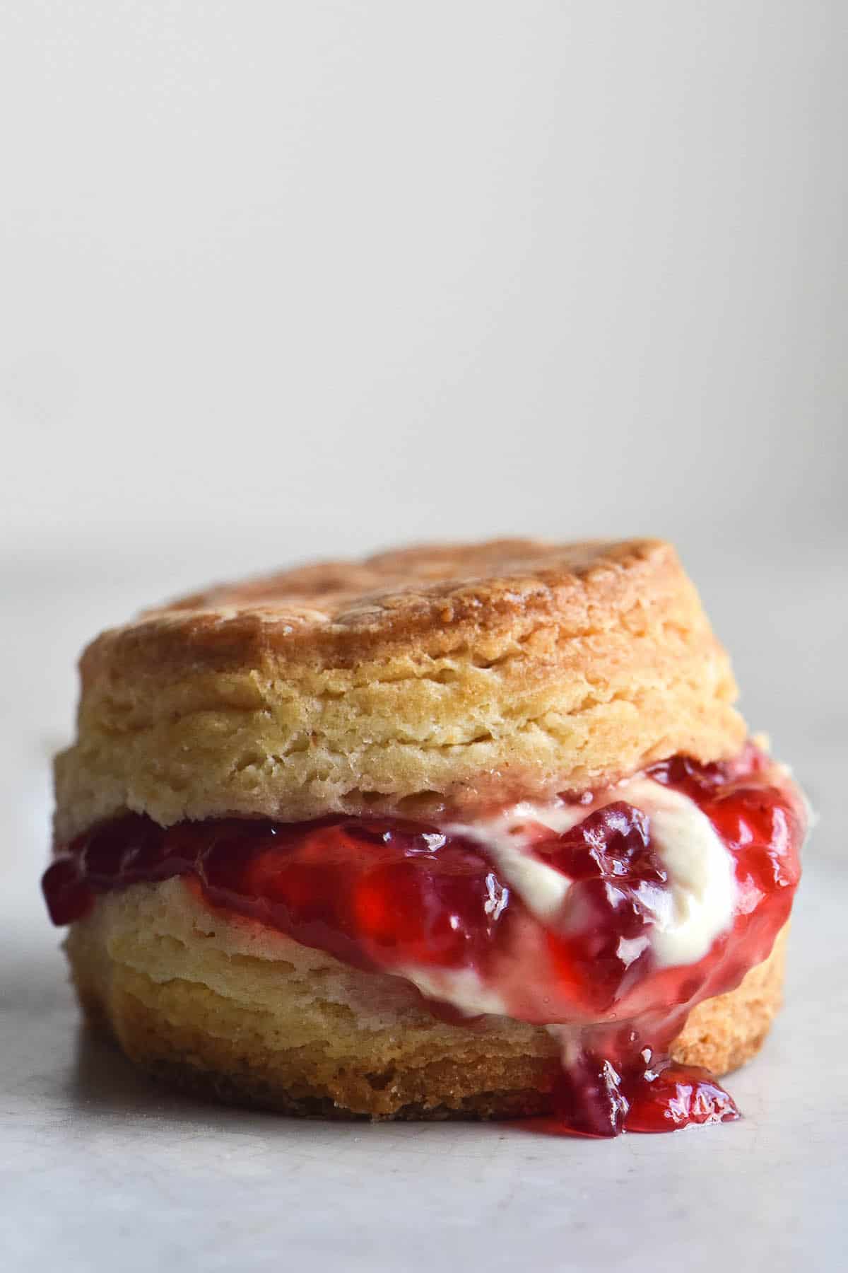 A side on image of a gluten free scone filled with jam and cream. The scone sits on a white marble table against a white backdrop