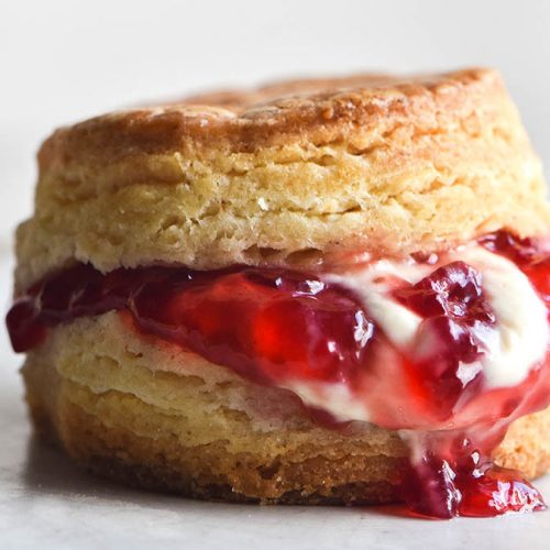 A close up side on view of a gluten free scone filled with jam and cream. The scone sits on a white ceramic plate against a white backdrop