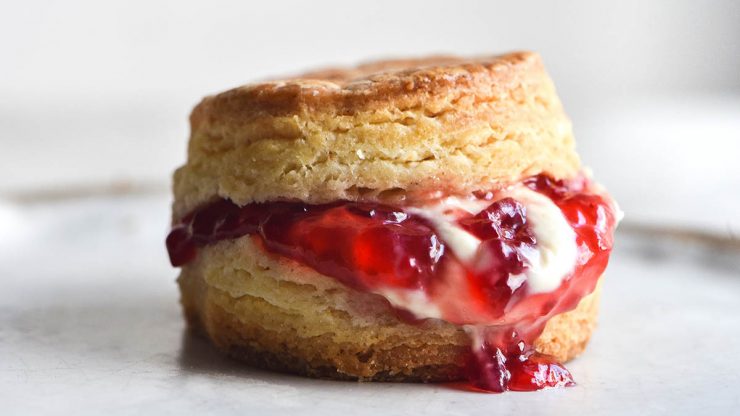 A close up side on view of a gluten free scone filled with jam and cream. The scone sits on a white ceramic plate against a white backdrop