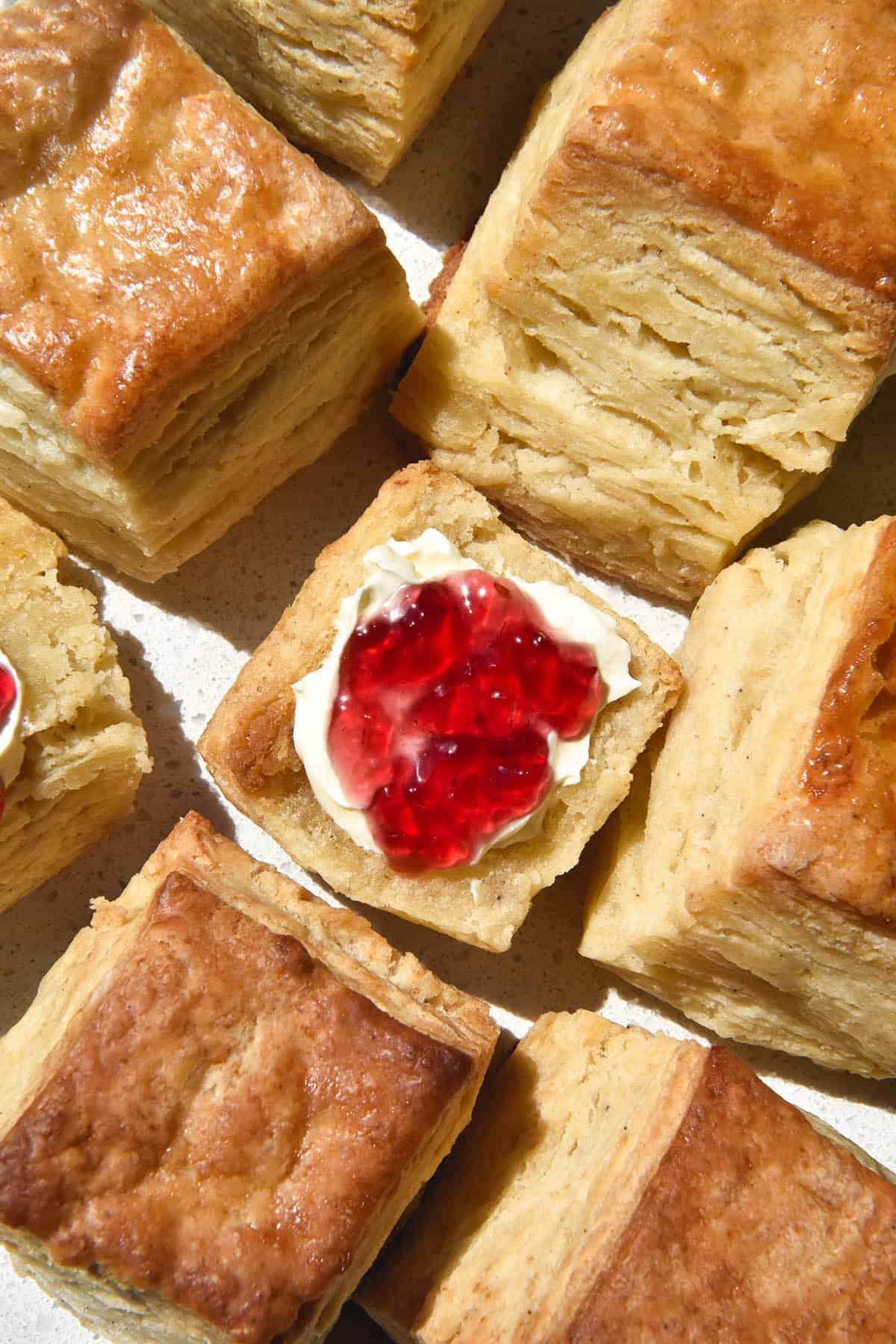 An aerial view of gluten free square scones on a white benchtop in bright sunlight. The centre scone has been halved and is topped with cream and jam