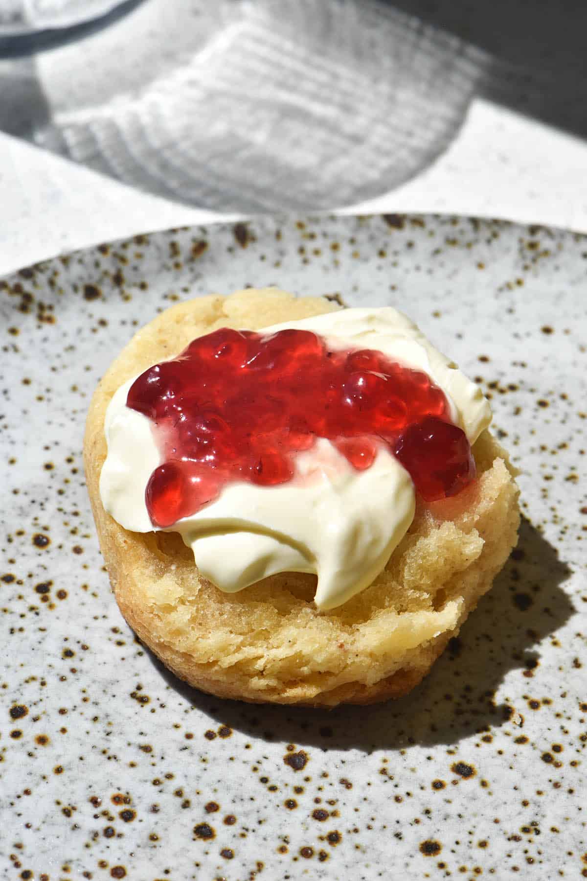 A side on view of a gluten free scone half topped with jam and cream. The scone sits on a white speckled ceramic plate and a glass of water sits in the background.