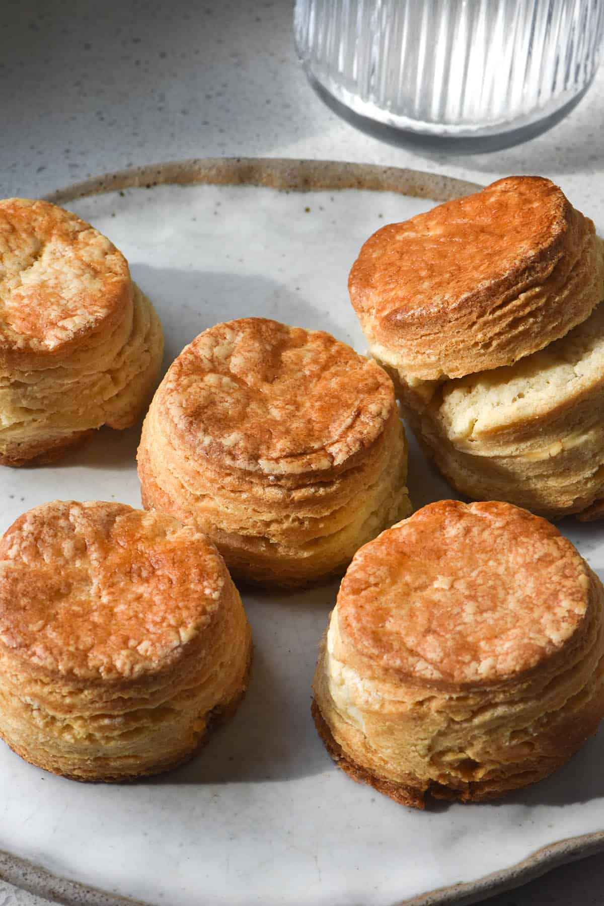 A white ceramic plate topped with flaky, layered and golden gluten free scones. The plate sits on a white stone benchtop and a textured glass sits in the top right corner