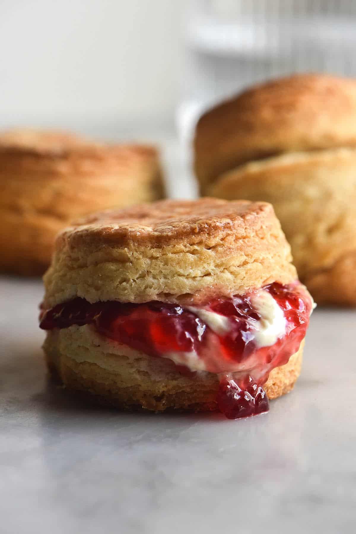 A side on image of a flaky gluten free scone filled with jam and cream. The scone sits on a white marble table and more scones sit in the background. 
