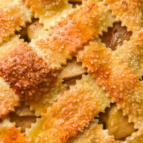 A close up macro image of the lattice pastry top of a gluten free apple pie. The lattice pastry is golden brown and topped with finishing sugar, and the apple filling pokes out between the lattice work.