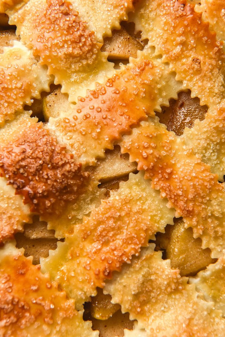 A close up macro image of the lattice pastry top of a gluten free apple pie. The lattice pastry is golden brown and topped with finishing sugar, and the apple filling pokes out between the lattice work.