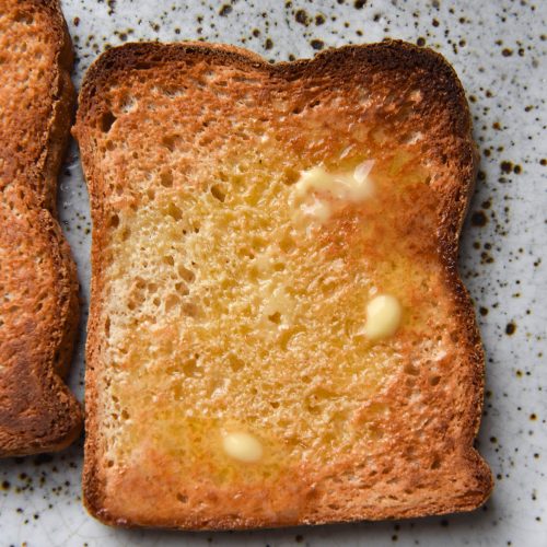 An aerial close up view of two pieces of toasted grain free bread on a white ceramic plate. The pieces of bread are golden brown and topped with pockets of melted butter