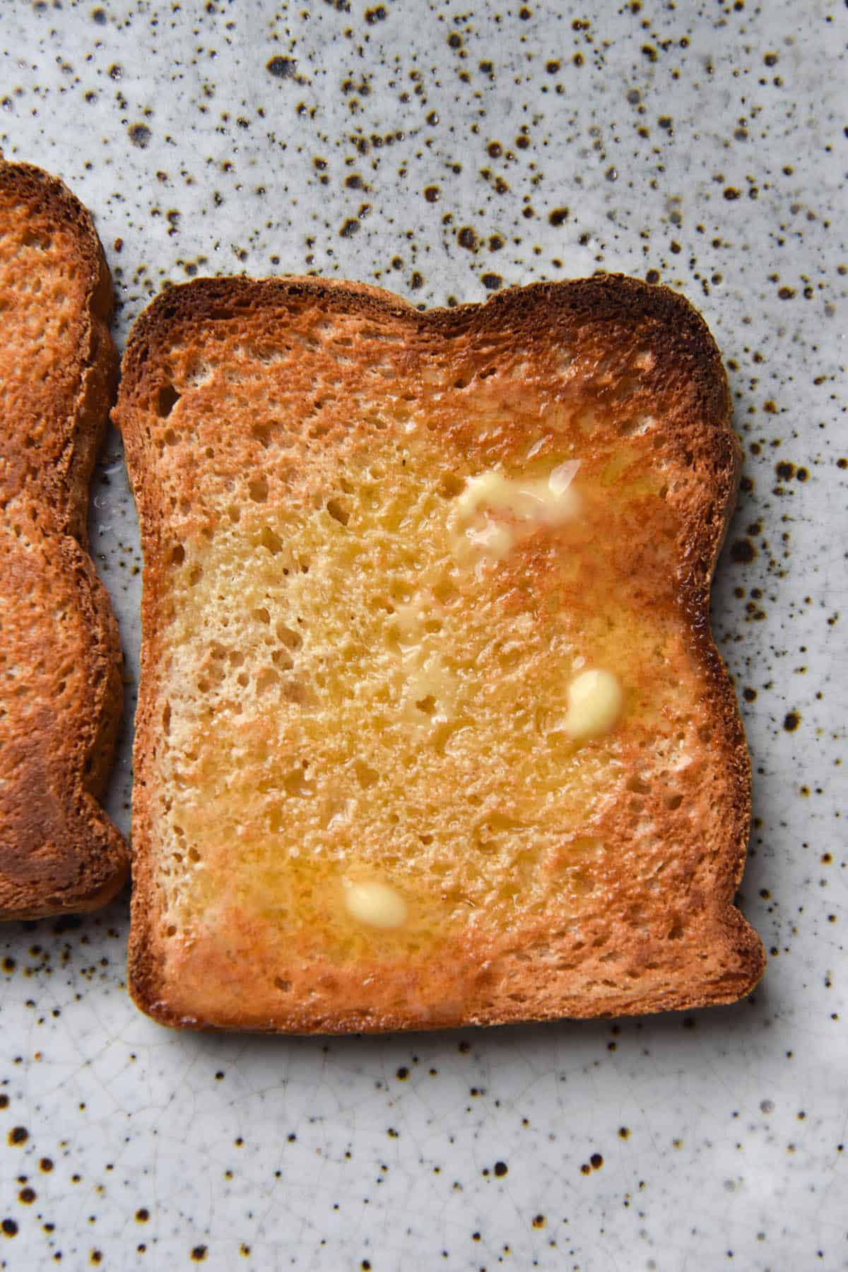 An aerial close up view of two pieces of toasted grain free bread on a white ceramic plate. The pieces of bread are golden brown and topped with pockets of melted butter