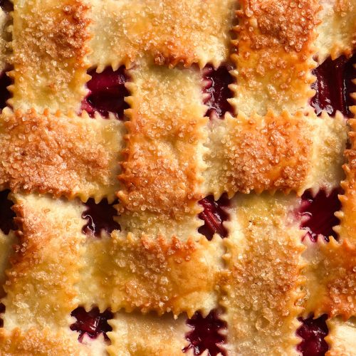 A close up macro photo of a gluten free strawberry pie finished with a lattice pastry stopping. The lattice pastry is topped with finishing sugar and has a golden brown appearance. Between the lattices, oozy strawberry peaks through.