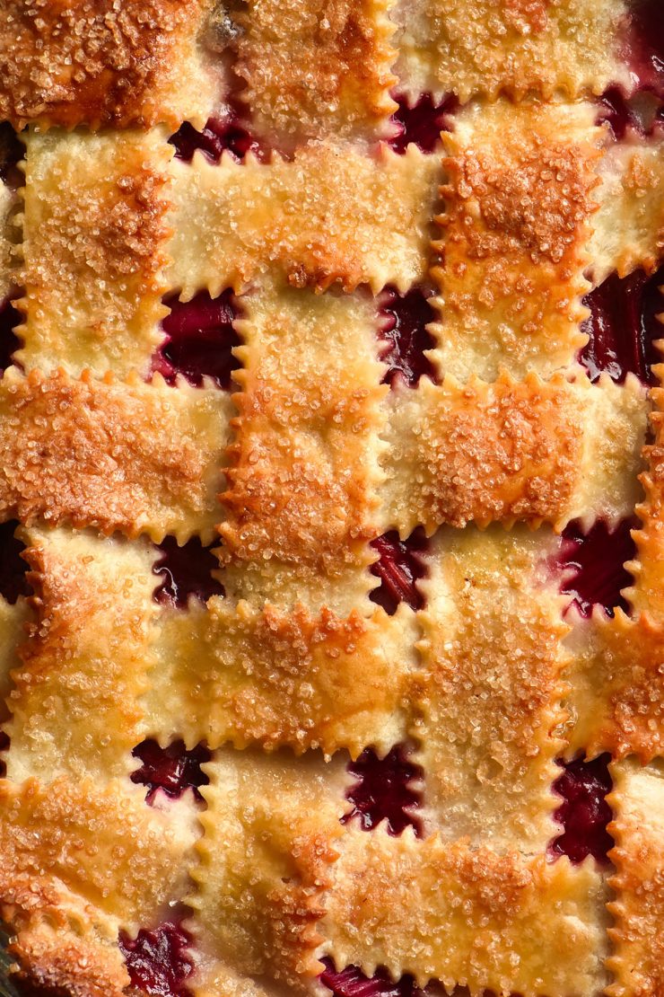 A close up macro photo of a gluten free strawberry pie finished with a lattice pastry stopping. The lattice pastry is topped with finishing sugar and has a golden brown appearance. Between the lattices, oozy strawberry peaks through.