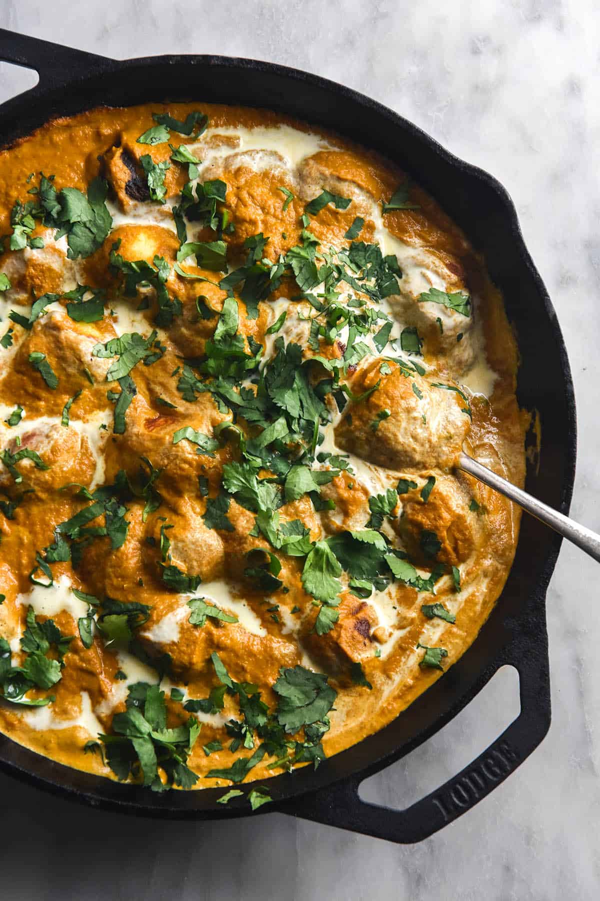 An aerial view of a skillet filled with FODMAP friendly Malai Kofta and topped with drizzled cream and chopped coriander. A spoon pokes out to the bottom left of the skillet, which sits on a white marble table
