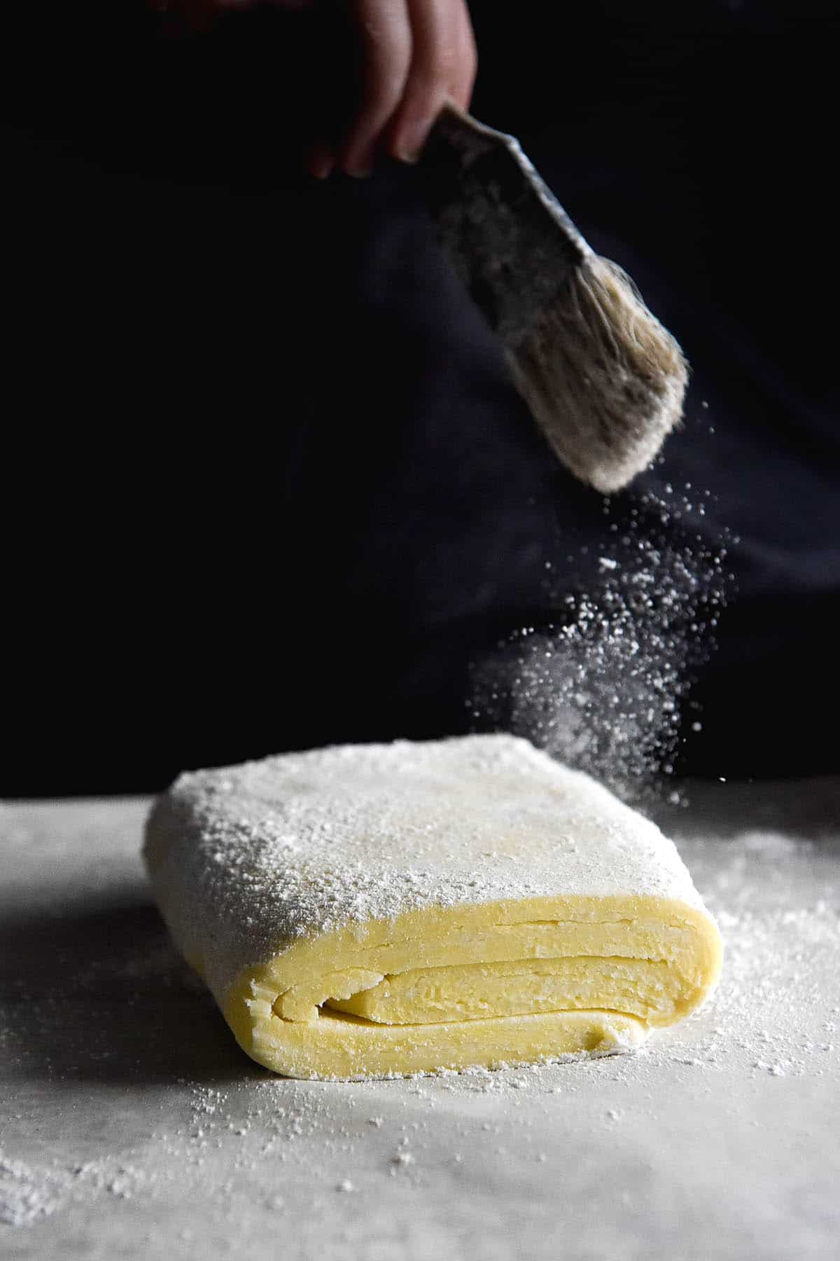 A slab of gluten free pastry sits on a piece of baking paper against a dark background. A hand extends from the top of the image to dust the excess flour off the pastry with a pastry brush. 