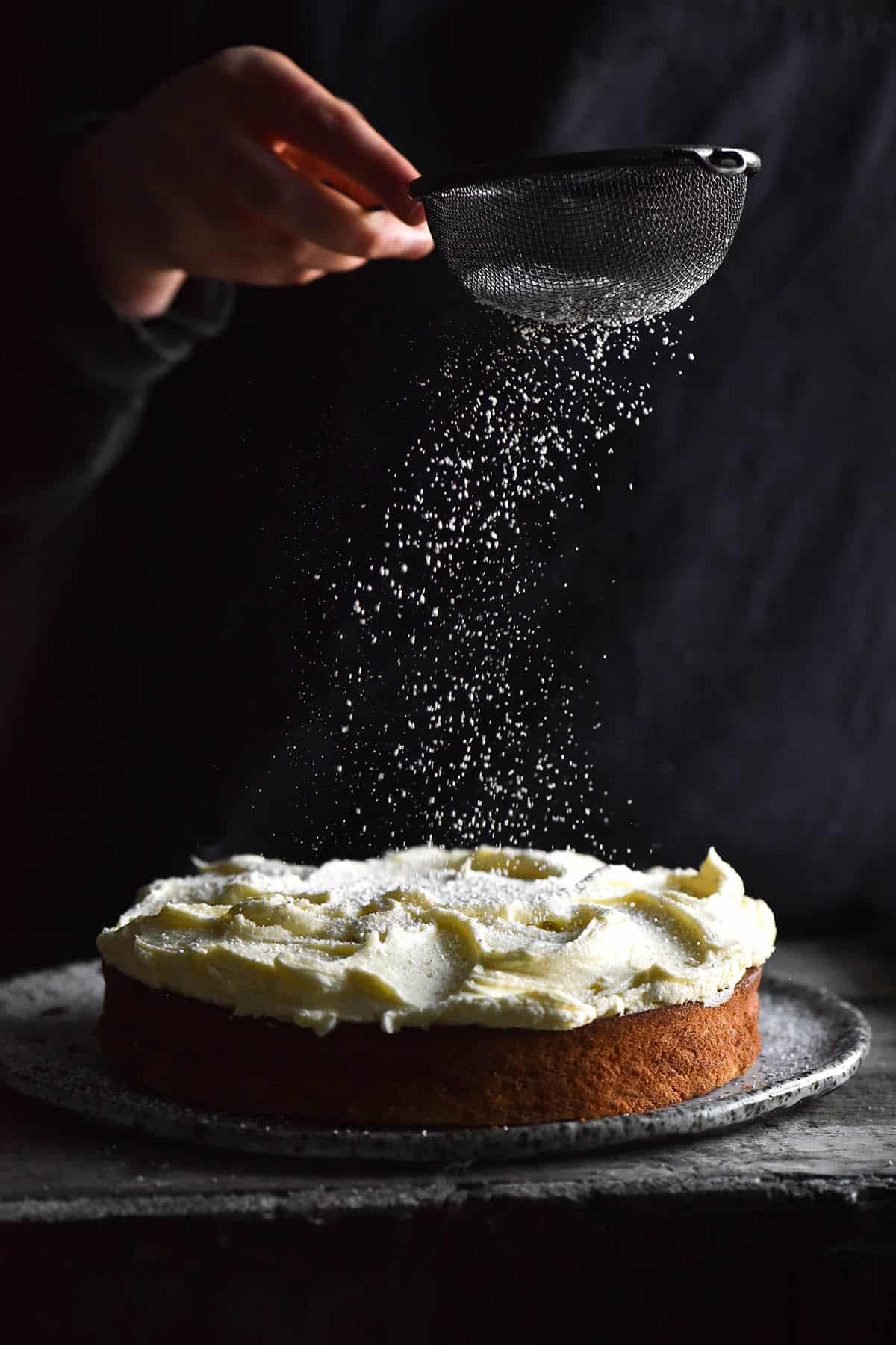 A side on moody image of a gluten free banana cake topped with cream cheese icing. The cake sits atop a white speckled ceramic plate against a dark grey backdrop. A hand extends from the right of the image to sprinkle icing sugar down onto the icing. The icing sugar is frozen in motion and contrasts with the dark background