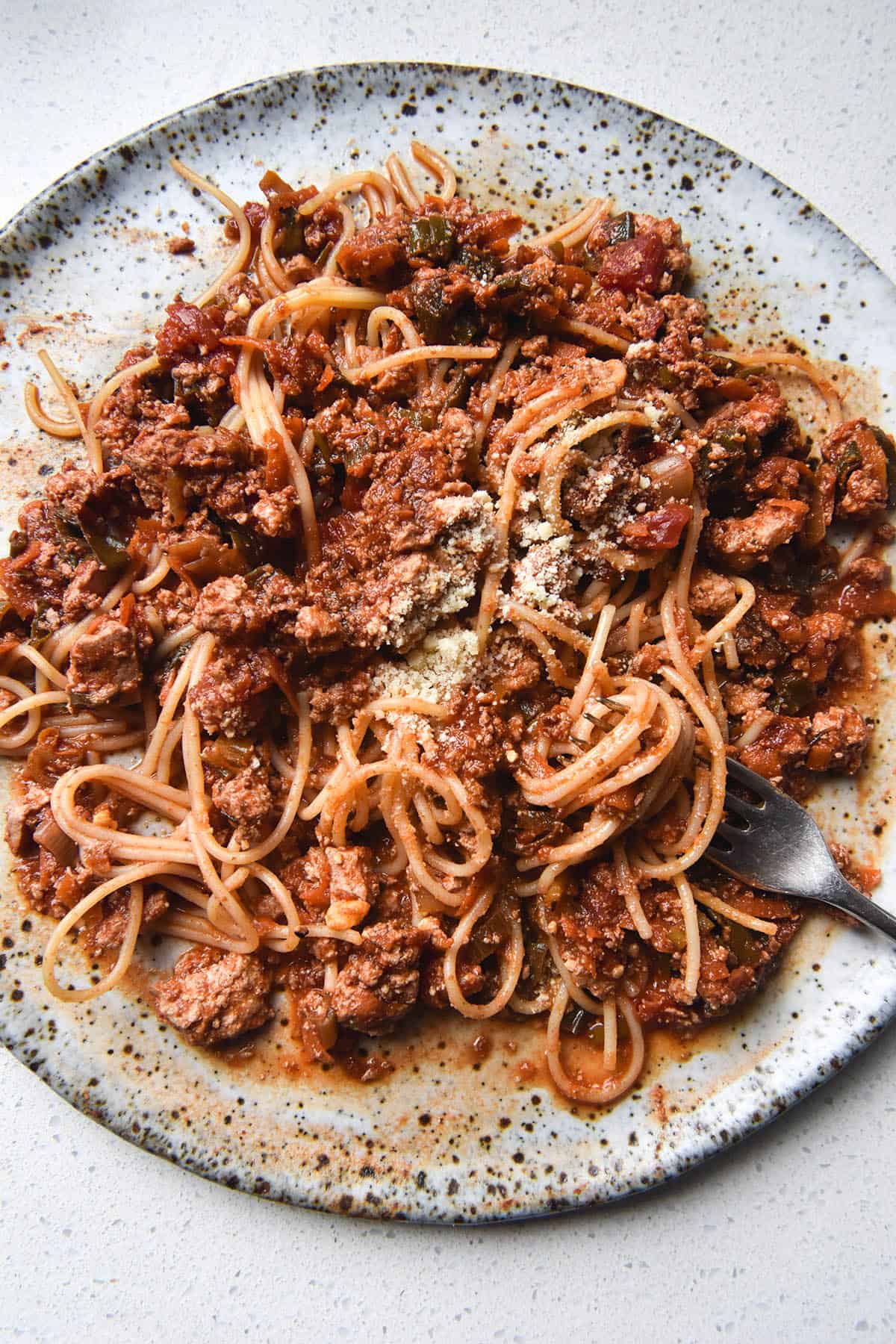 An aerial view of a white speckled ceramic plate topped with vegan spaghetti bolognese. A fork sticks into the bolognese from the bottom right of the image