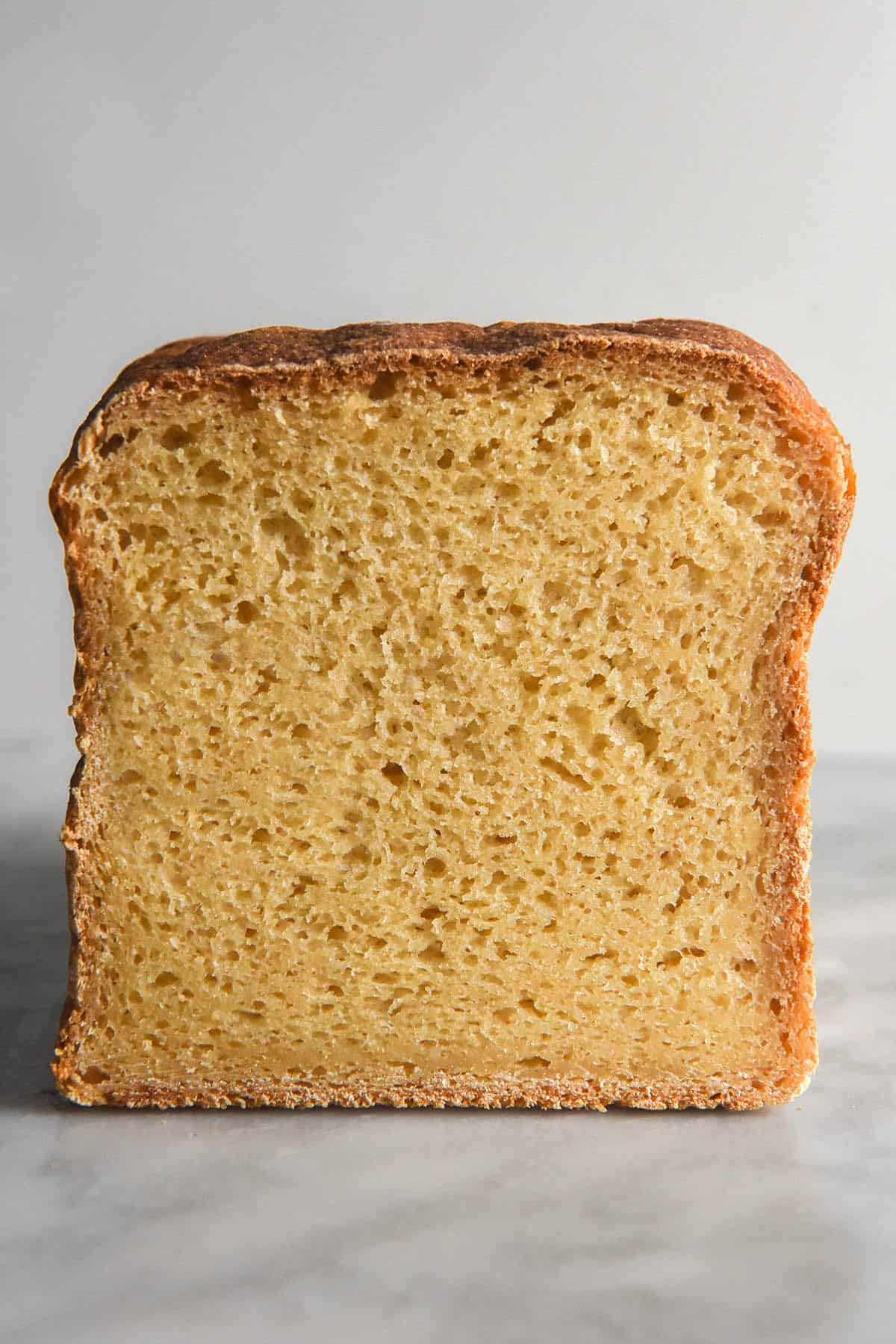 A close up side on view of a loaf of grain free bread on white table against a white backdrop. A slice has been cut from the loaf, revealing the fluffy crumb underneath