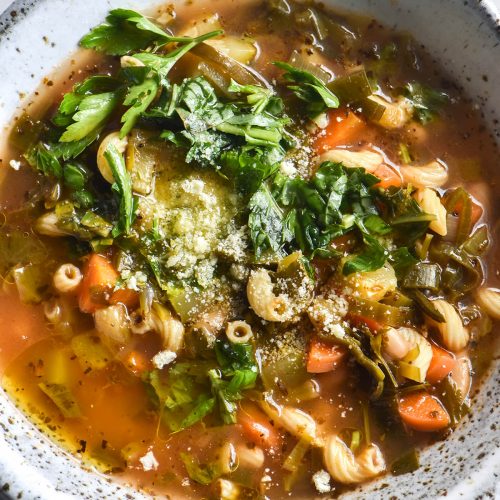 An aerial close up view of a white speckled ceramic bowl filled with low FODMAP minestrone. The minestrone is topped with garlic infused oil, fresh herbs and parmesan. The bowl sits atop a white marble table