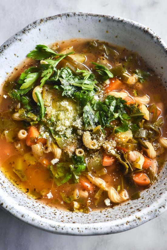 An aerial close up view of a white speckled ceramic bowl filled with low FODMAP minestrone. The minestrone is topped with garlic infused oil, fresh herbs and parmesan. The bowl sits atop a white marble table