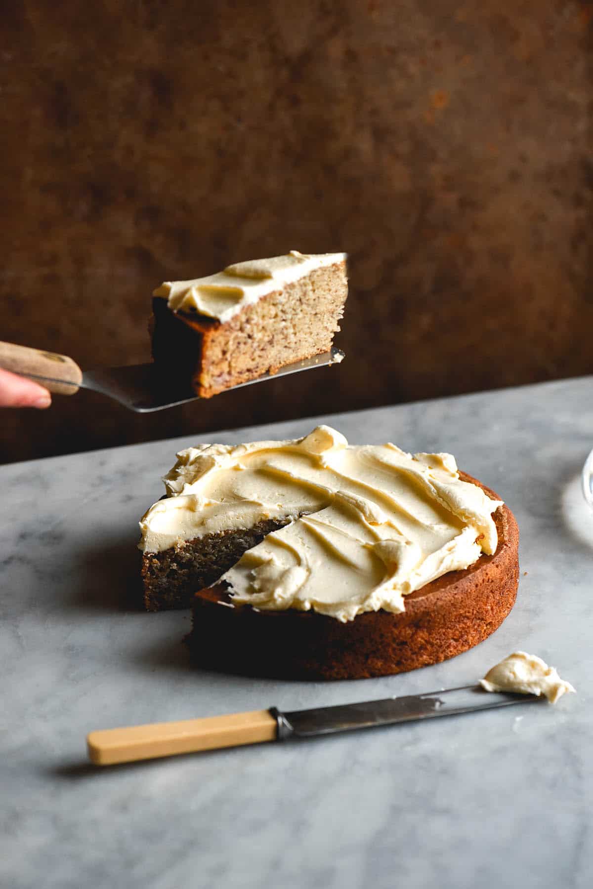 A side on view of a gluten free banana cake on a white marble table against a rusty orange backdrop. The cake is topped with cream cheese icing and a slice has been removed. A hand extends from the left of the image to lift the cake slice up from the cake. 