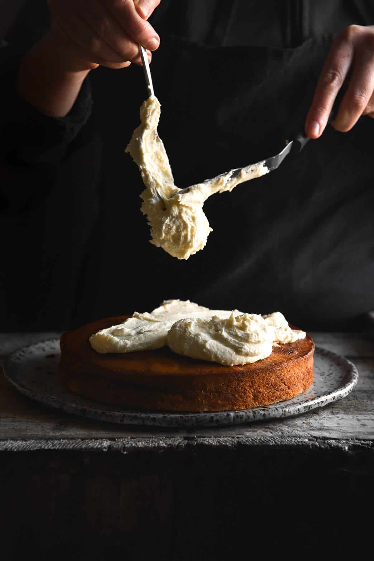 A moody side on image of a gluten free banana cake on a dark wooden bench. A person stands behind the cake in a black apron and jumper to spoon cream cheese icing down onto the cake. 
