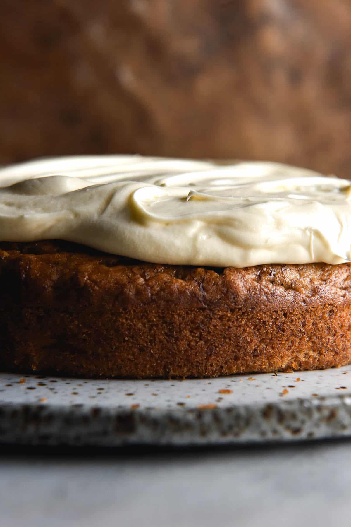 A side on close up image of a gluten free banana cake topped with plenty of fluffy cream cheese icing. The cake sits on a white speckled ceramic plate atop a white marble table. The background is a rusty auburn steel backdrop. 