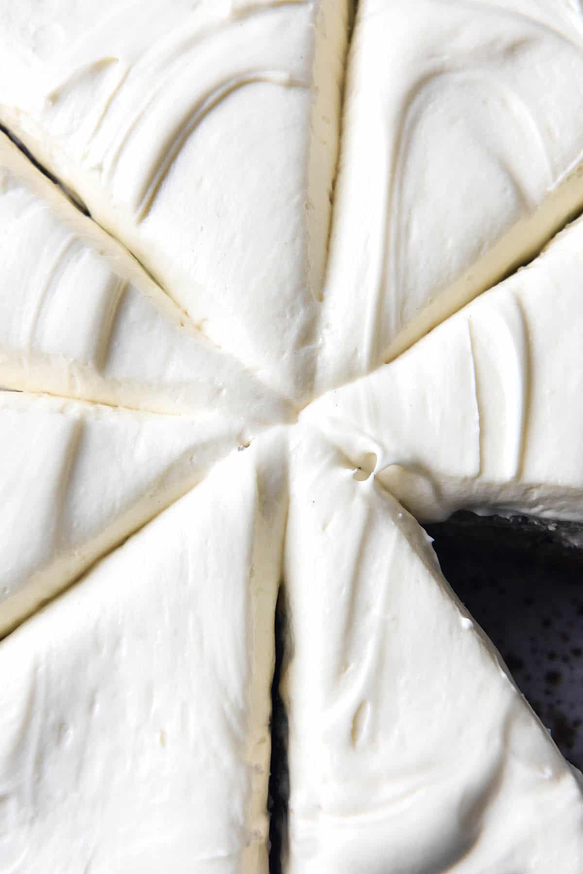 A close up macro image of a gluten free banana cake topped with fluffy cream cheese icing. The cake has been cut into 8, making a slice pattern through the top of the white icing. The bottom right slice of cake has been removed, revealing a speckled ceramic plate underneath. 