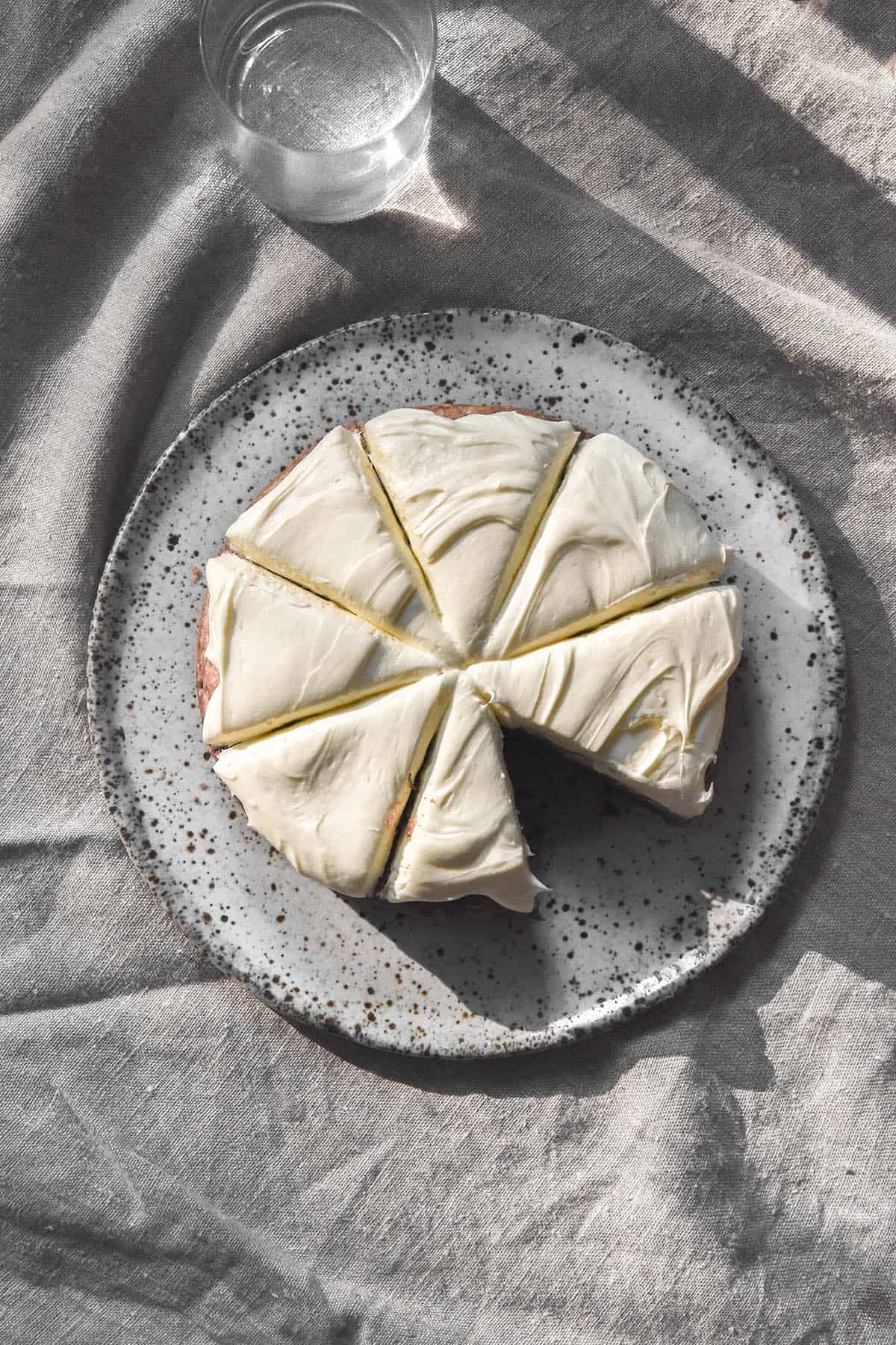 An aerial view of a gluten free banana cake topped with cream cheese icing. The cake sits on a white speckled ceramic plate against a beige linen tablecloth. The cake is sliced into eight pieces and the bottom right piece has been removed. 