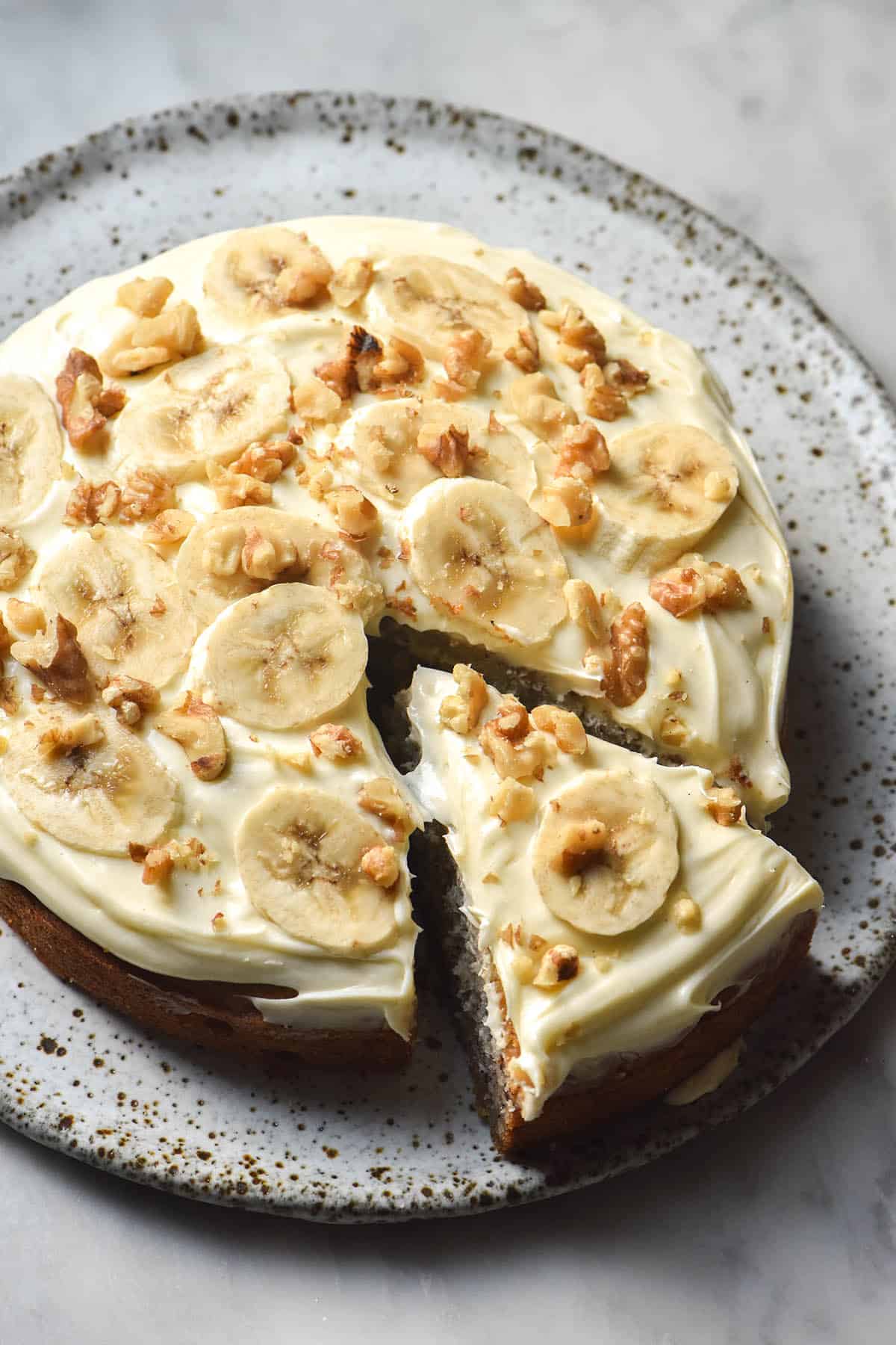 An aerial view of a gluten free banana cake on a white speckled plate atop a white marble table. The banana cake is topped with cream cheese icing, sliced bananas and crushed walnuts