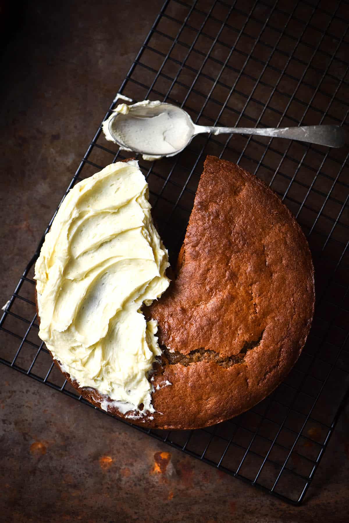 An aerial view of a gluten free banana cake on a rusty auburn coloured backdrop. The cake is half covered in cream cheese icing and half plain. A slice has been removed from the top of the cake, and a spoon covered in cream cheese sits above the cake.