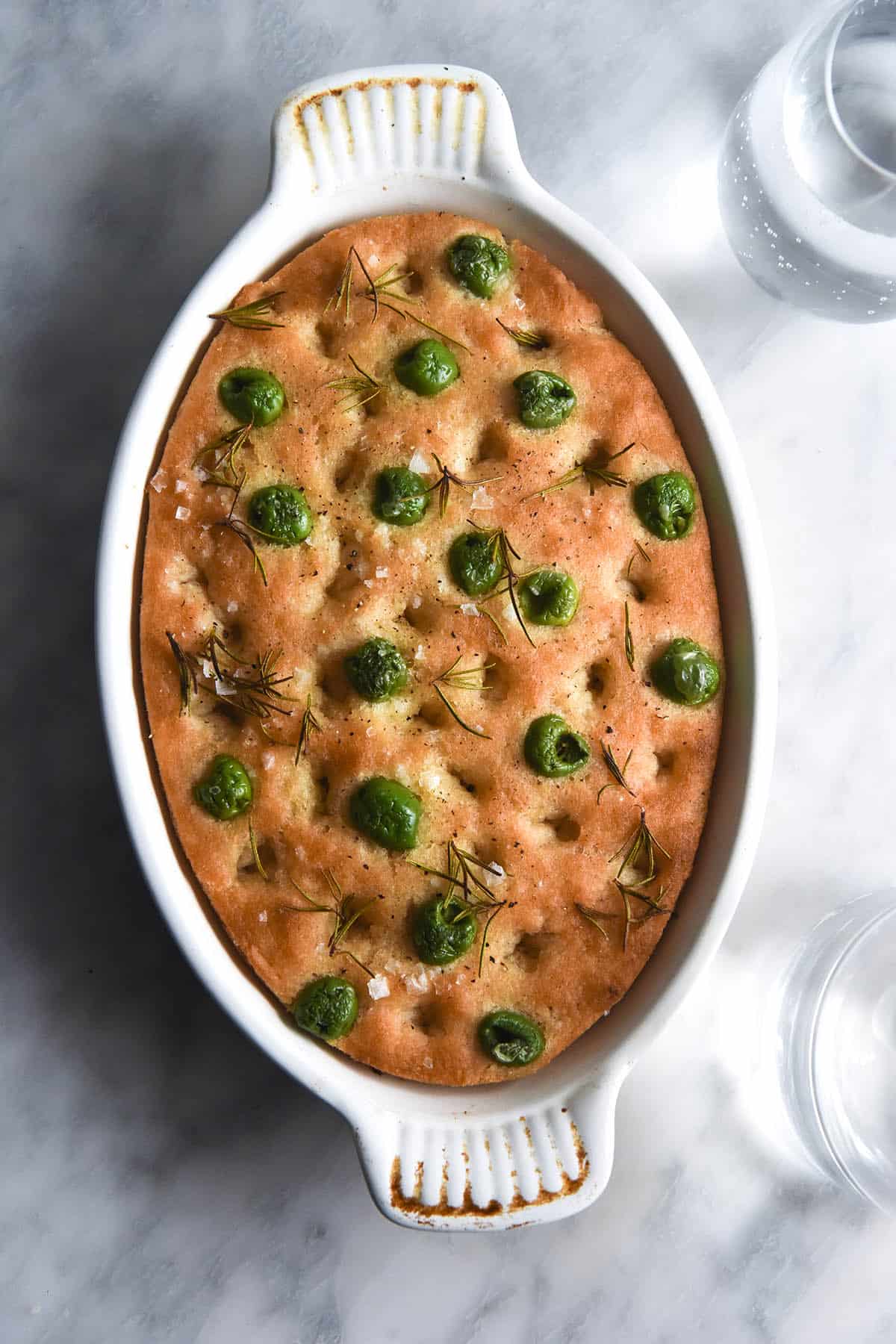 An aerial image of a gluten free focaccia in a white oval shaped baking dish on a white marble table. The focaccia is topped with olives, rosemary sprigs and sea salt flakes. 