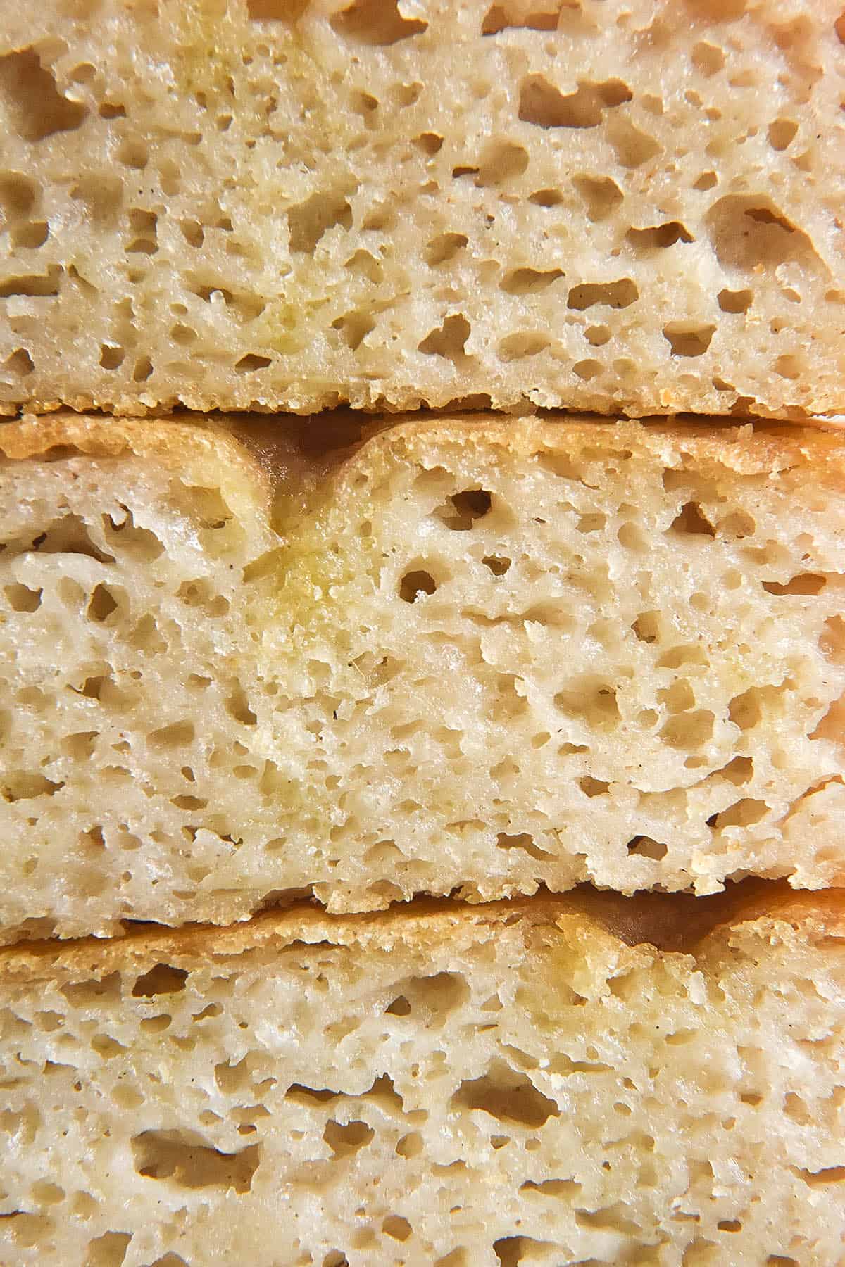 A side on close up macro image of a stack of gluten free focaccia slices. The focaccia crumb is exposed, revealing a wide open crumb and olive oil oozing throughout the focaccia