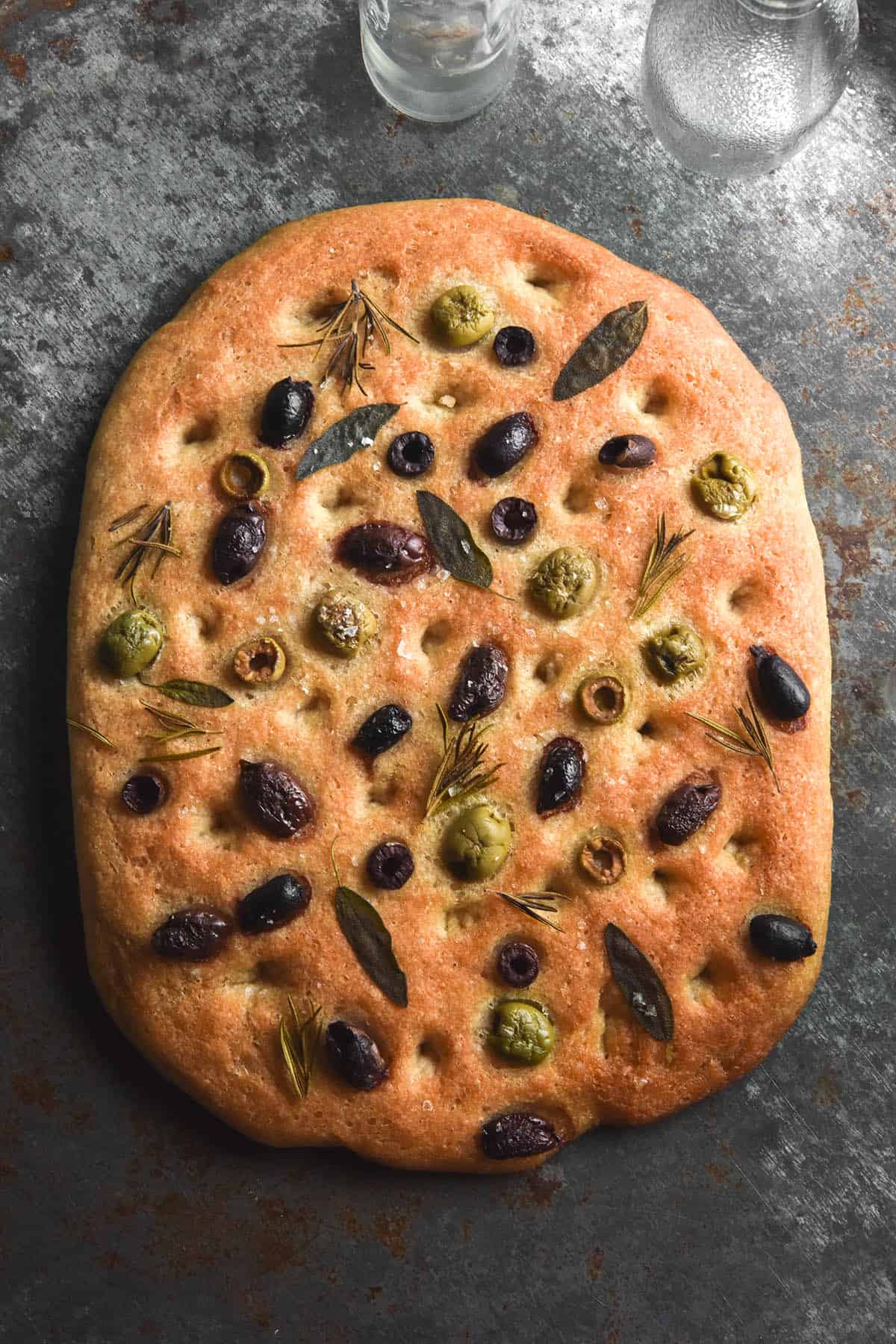 An aerial image of a gluten free focaccia on a dark steel background. The focaccia is golden brown and topped with green and kalamata olives, sage leaves, rosemary leaves and sea salt flakes. 