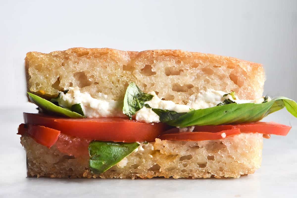 A side on image of a gluten free focaccia sandwich on a white marble table against a white backdrop. The sandwich is filled with tomato, basil and vegan mozzarella, which pokes out of the side of the sandwich. 