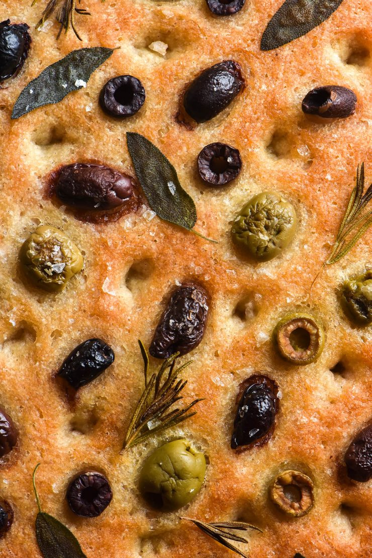 A close up aerial image of a gluten free focaccia that is golden brown and topped with green olives, kalamata olives, sage leaves, rosemary sprigs and flaky sea salt