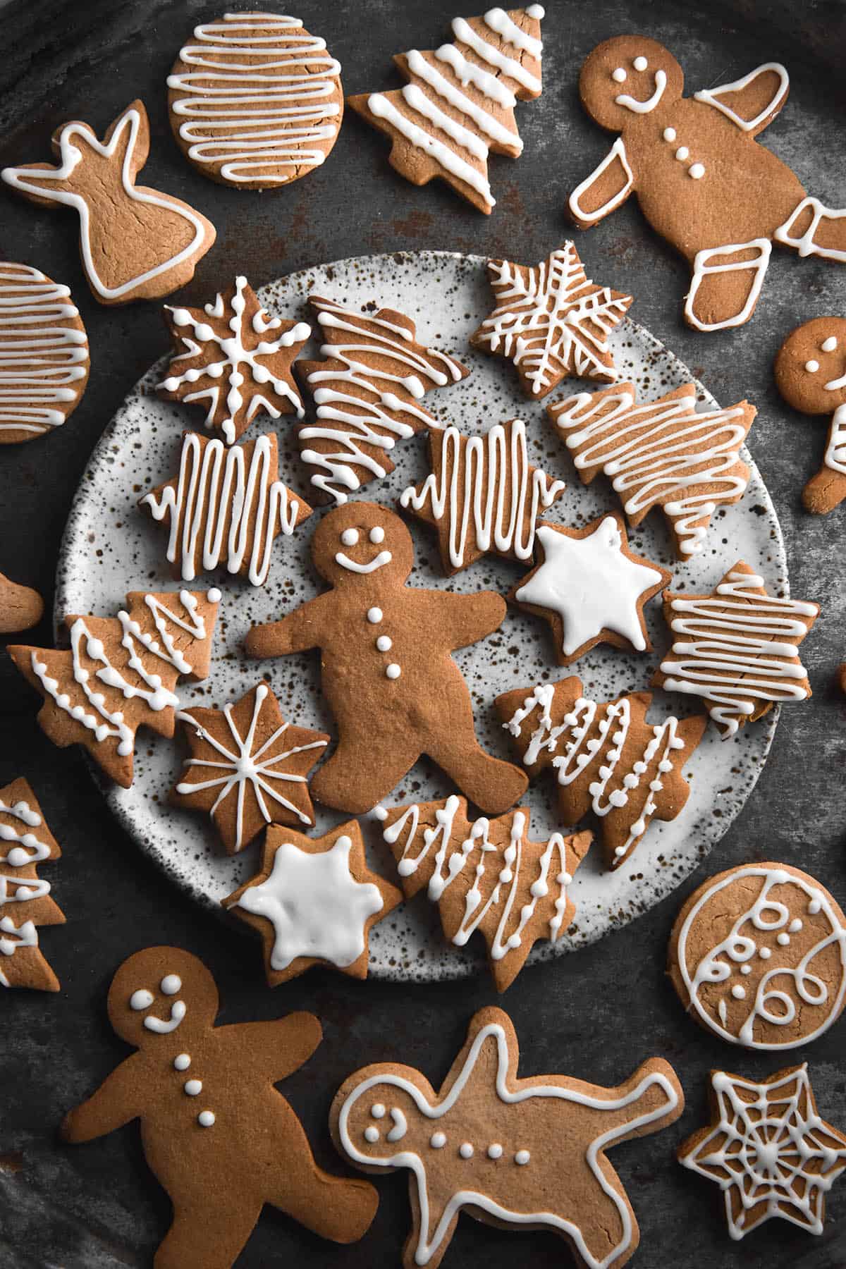 An aerial image of gluten free gingerbread cookies decorated with white icing on a dark steel backdrop. A white ceramic speckled plate sits in the centre of the image, topped by more gingerbread. 