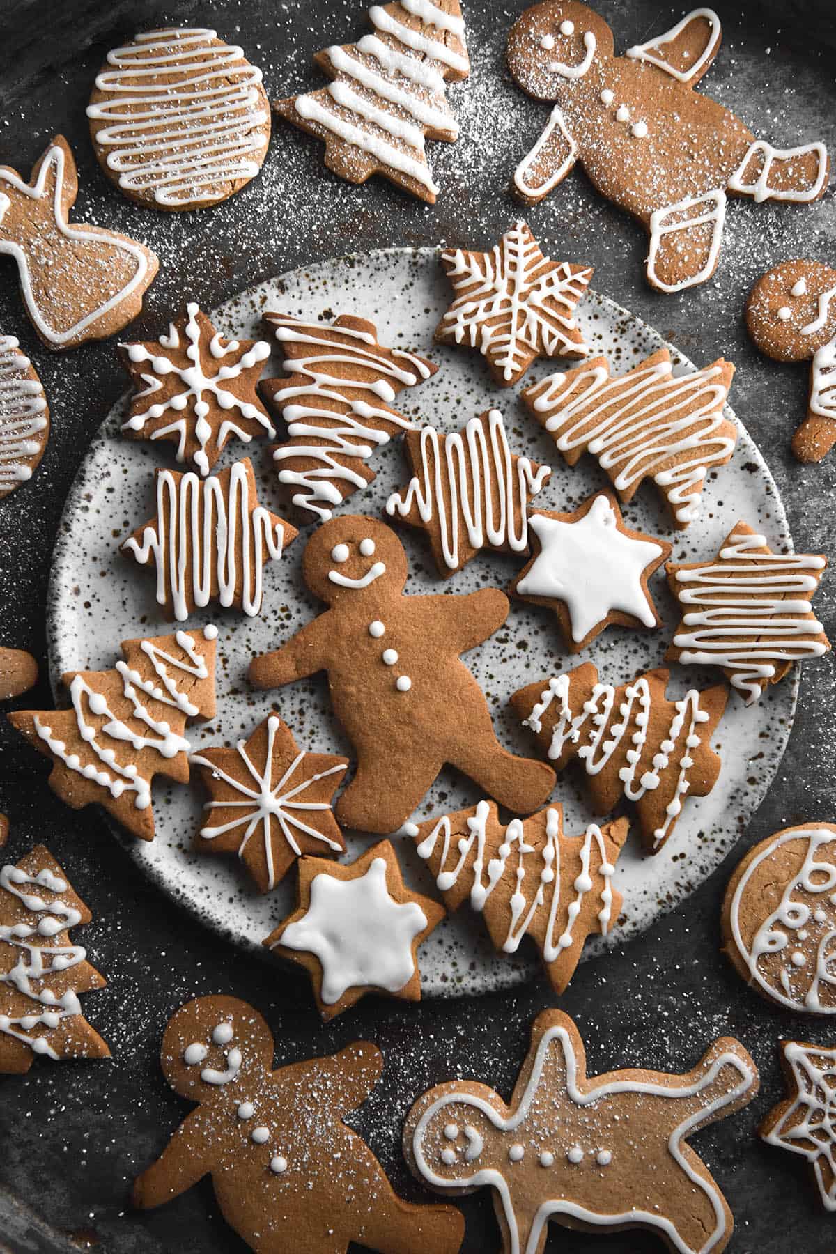 An aerial image of a gluten free gingerbreads decorated with white icing. The gingerbread around the edge of the image sit atop a dark steel backdrop and are sprinkled by extra icing sugar. A white speckled ceramic plate sits in the middle of the image topped with the remaining gingerbread