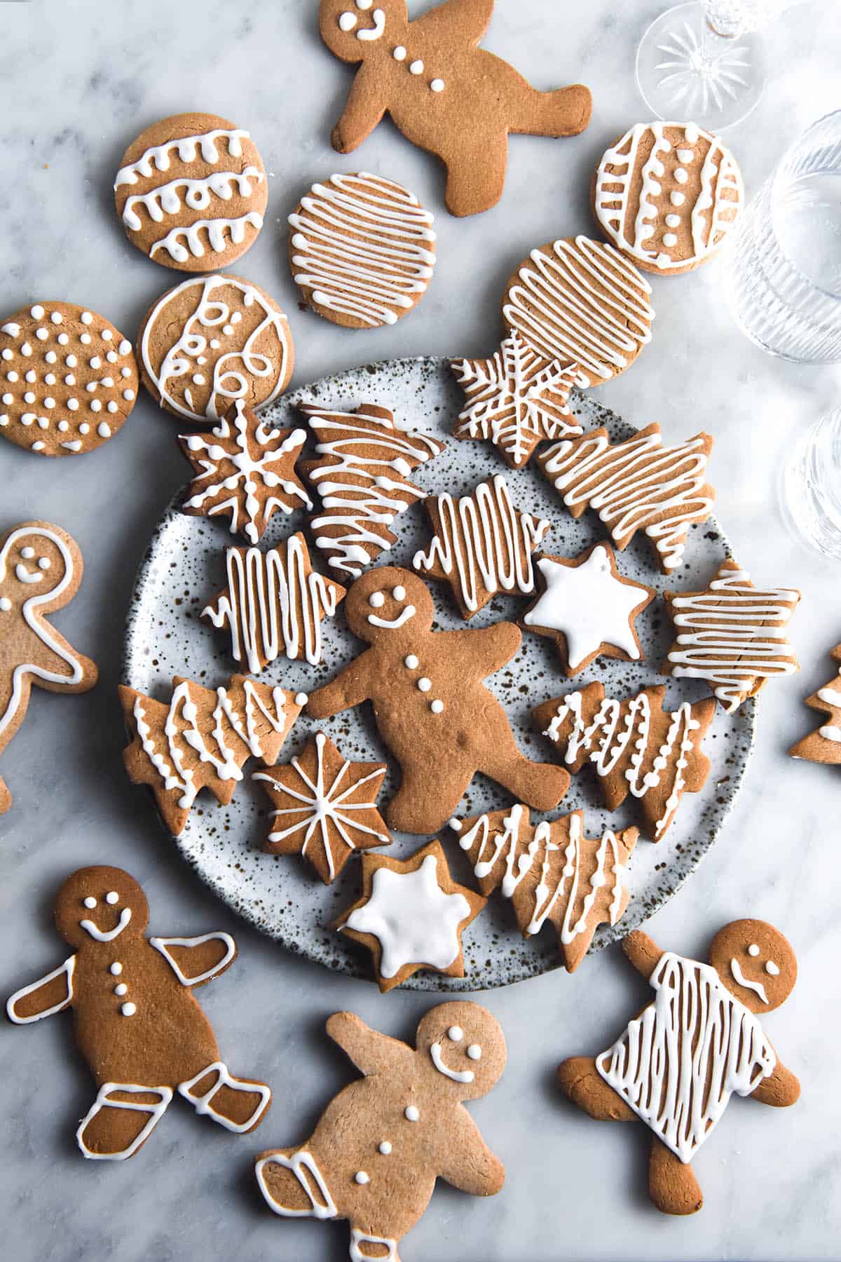 An aerial view of a white marble table casually strewn with gluten free gingerbread cookies in various shapes. The majority of the cookies are in the centre of the image on a white speckled ceramic plate. The cookies are decorated with white icing in various patterns