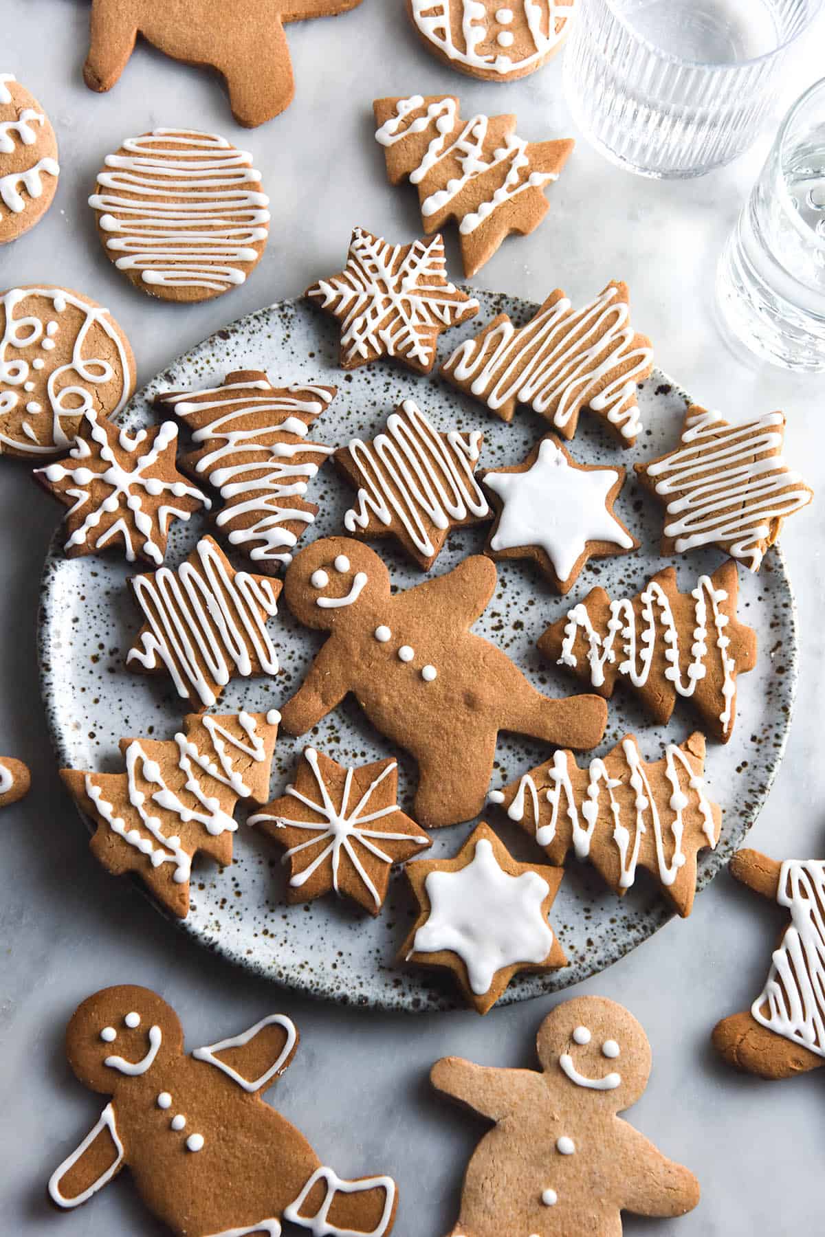 An aerial view of a white speckled ceramic plate topped with gluten free gingerbread cookies decorated in white icing with various patterns.