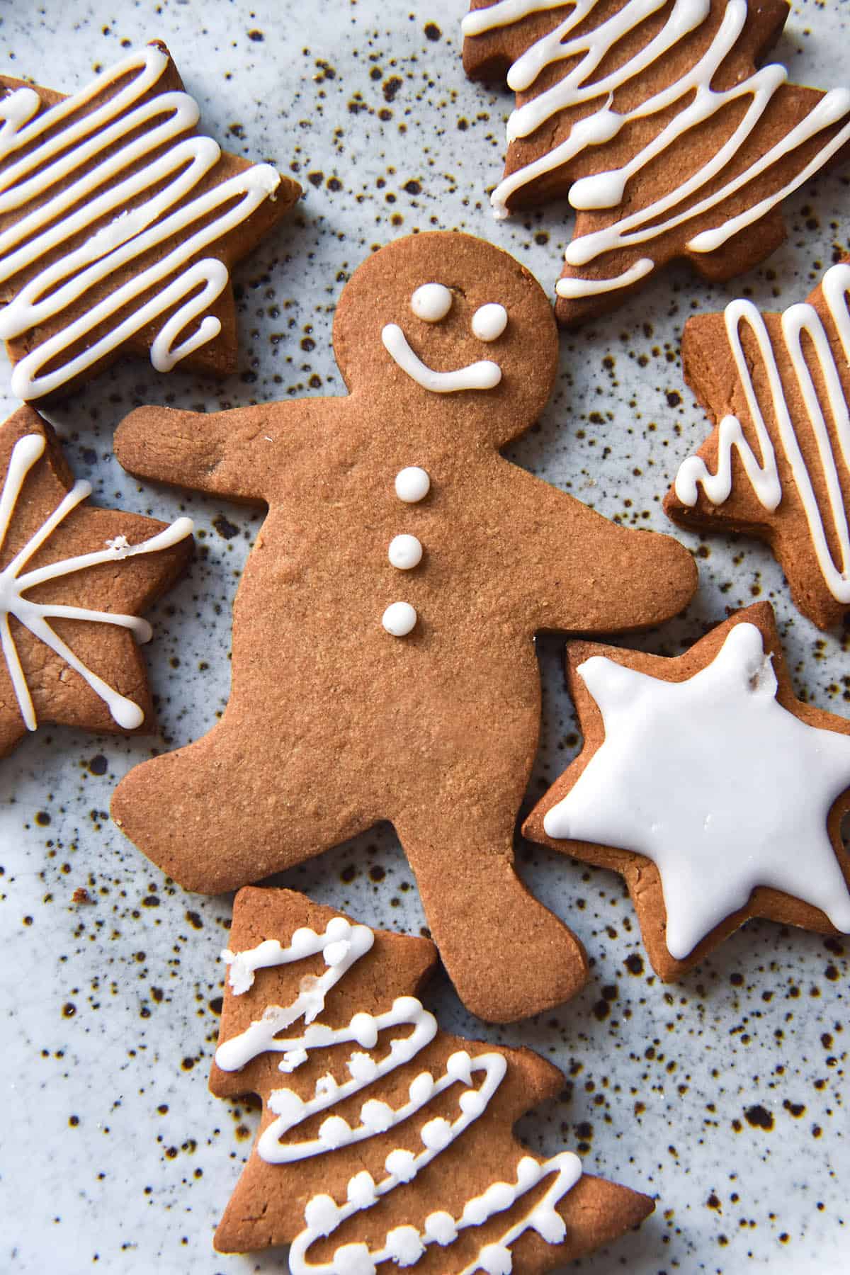 An aerial close up view of gluten free gingerbreads on a white speckled ceramic plate. The cookies are topped with white icing in various decorative patterns. 