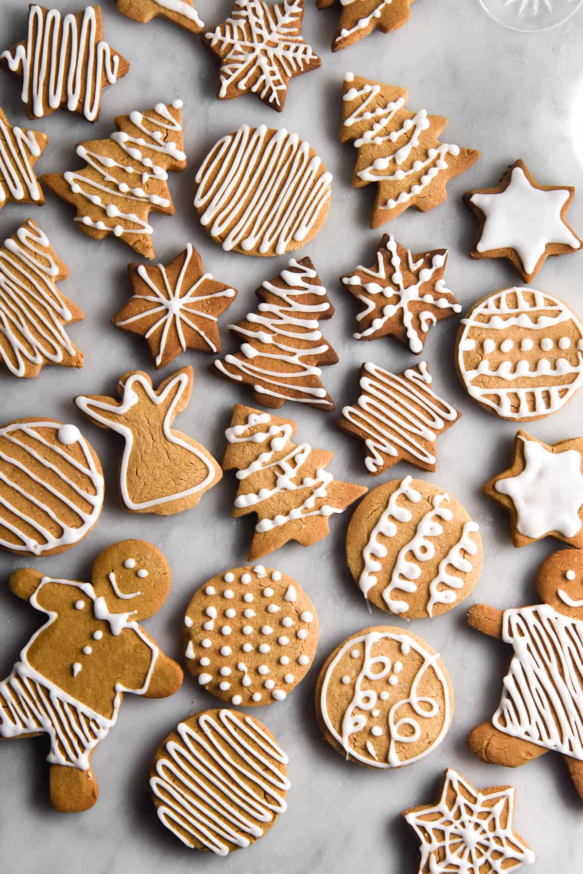 An aerial view of gluten free and vegan gingerbread cookies cut into various shapes and decorated with white icing sugar. The cookies are casually arranged on a white marble table.