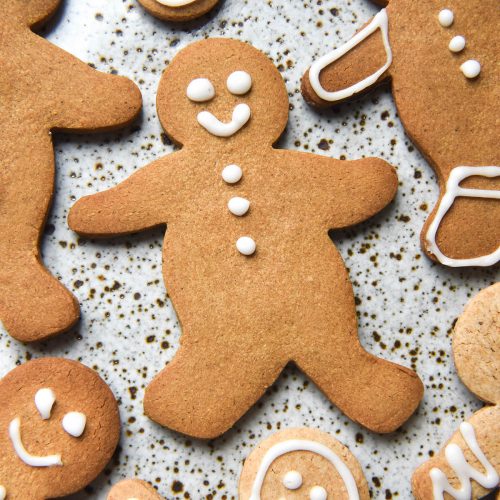 An aerial view of gluten free gingerbread people atop a white speckled ceramic plate. The gingerbread are lightly decorated with white icing, giving them smiley faces and other small details