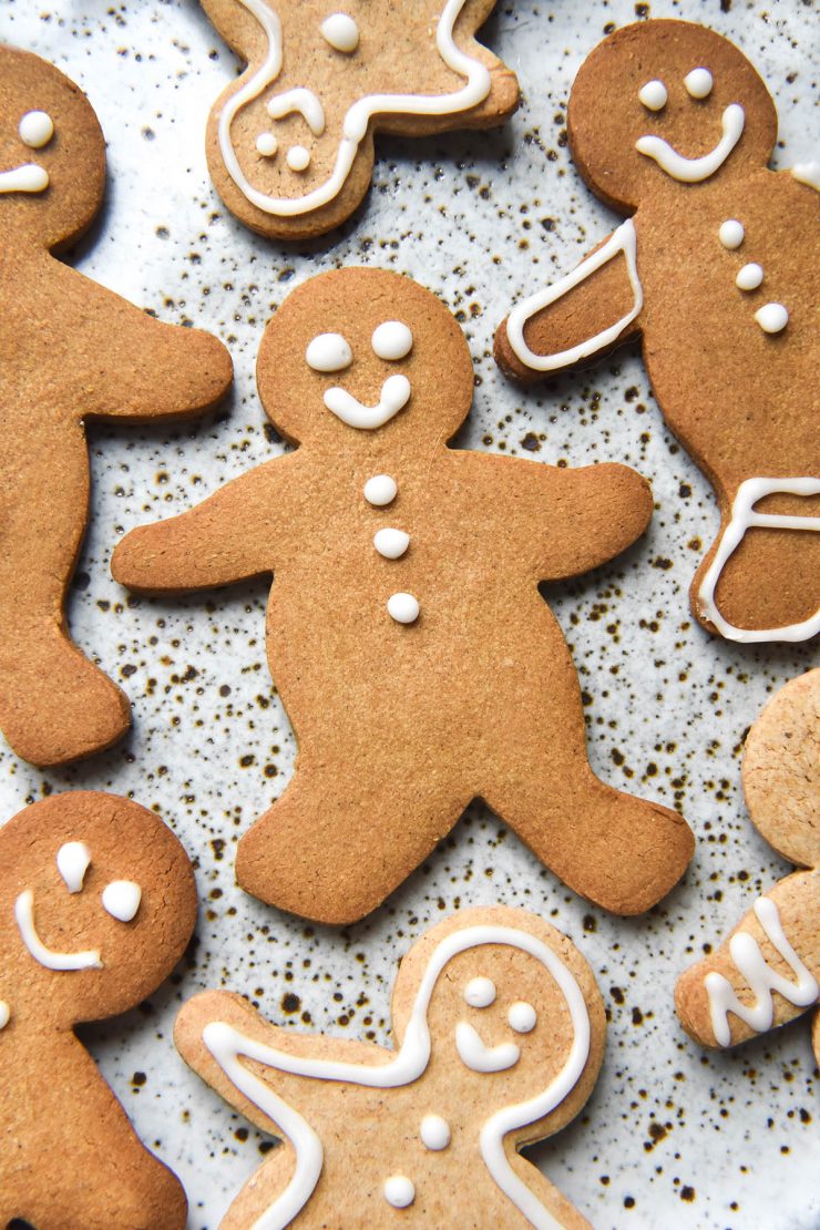 An aerial view of gluten free gingerbread people atop a white speckled ceramic plate. The gingerbread are lightly decorated with white icing, giving them smiley faces and other small details